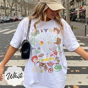 Pode incluir: T-shirt branco com um gr&aacute;fico colorido com personagens de desenhos animados e o texto "REACH FOR THE SKY". O design inclui um cacto, uma bota de cowboy e outros elementos l&uacute;dicos. A camisa tem um ajuste casual e oversized.