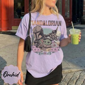 May include: A light purple t-shirt featuring a graphic of The Mandalorian, Grogu, and a spaceship, surrounded by flowers. The word "MANDALORIAN" is printed across the top. The shirt is paired with a black skirt.