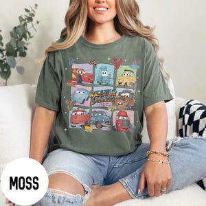 May include: Olive green t-shirt featuring a grid of cartoon cars from the movie Cars. The shirt has the text "Racing Springs" in a stylized font. The word "Moss" is printed on a white circle in the lower left corner.