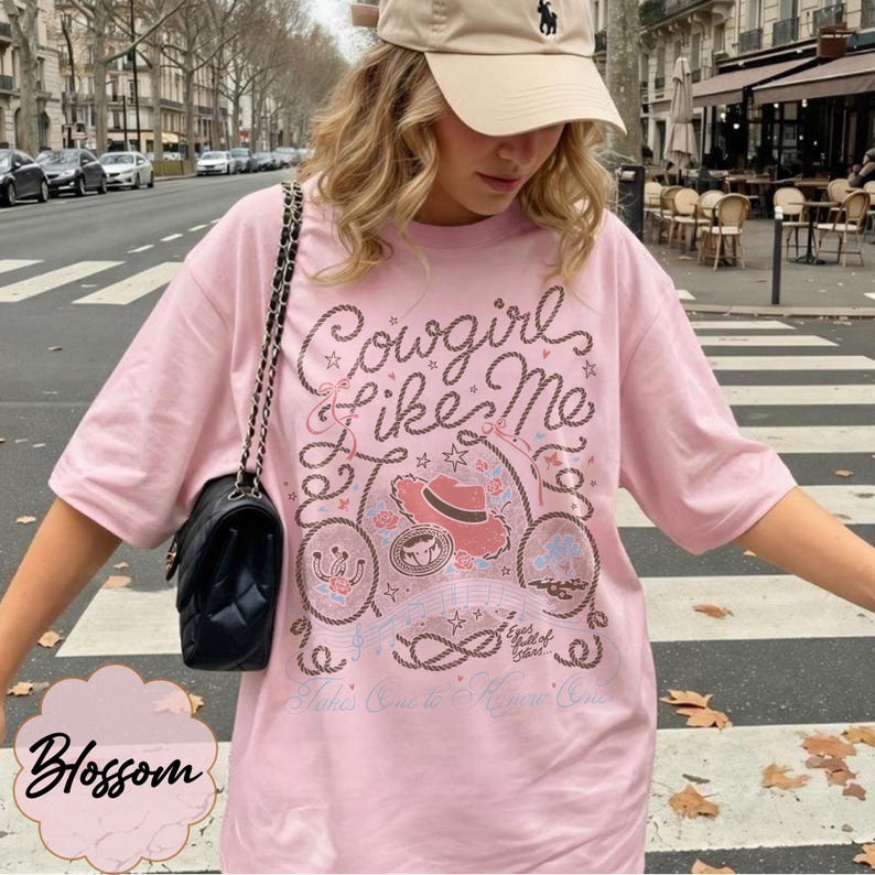 May include: A light pink short-sleeve t-shirt featuring a graphic print with the text Cowgirl Like Me, Eyes Full of Stars, and Takes One to Know One. The design includes a cowboy hat, rope motifs, stars, and floral elements in brown and red.