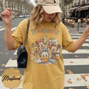 Pu&ograve; includere: T-shirt giallo senape con grafica Disneyworld 2026 con Topolino, Minnie, Paperino, Pippo e Pluto. La maglietta ha girocollo e maniche corte. La parola "Mustard" &egrave; scritta in corsivo.