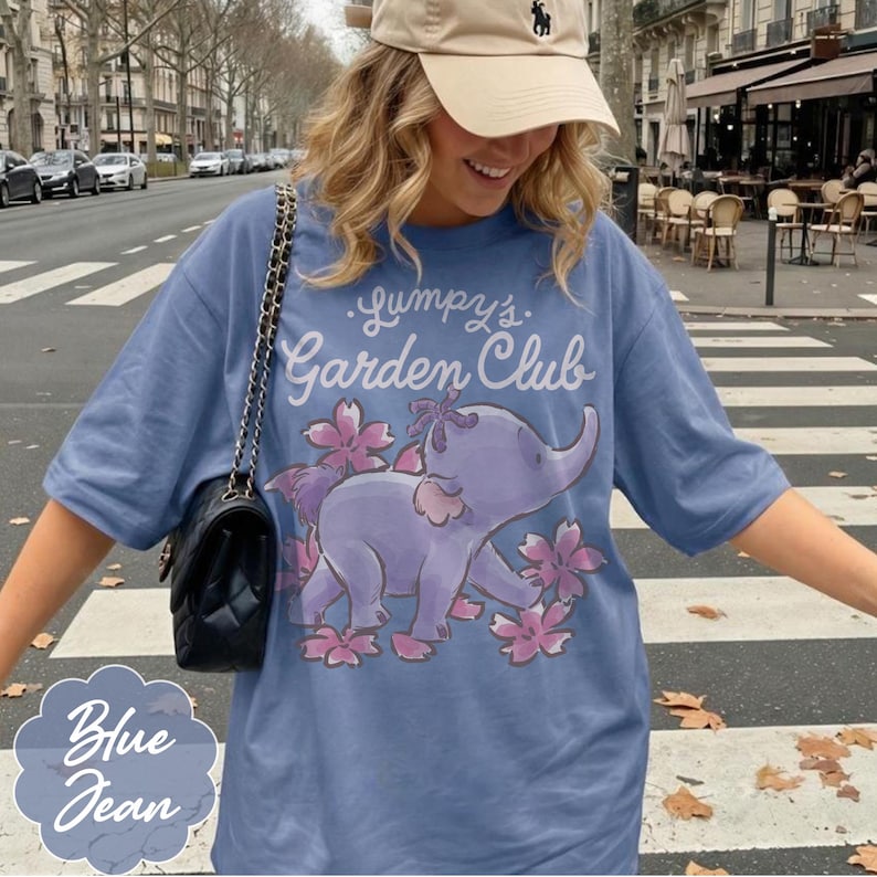 Op de afbeelding: Blauw spijkerstof T-shirt met een illustratie van een paarse olifant omringd door roze bloemen. De tekst "Gumpy's Garden Club" staat boven de olifant. Een lichtbruine baseballpet en een zwarte handtas zijn ook zichtbaar.