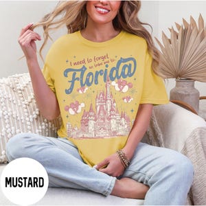 May include: Mustard-colored t-shirt with the text "I need to forget so take me to Florida" in blue script. The shirt features a castle graphic with pink and white accents, and Mickey Mouse-shaped clouds. The shirt is a relaxed fit.
