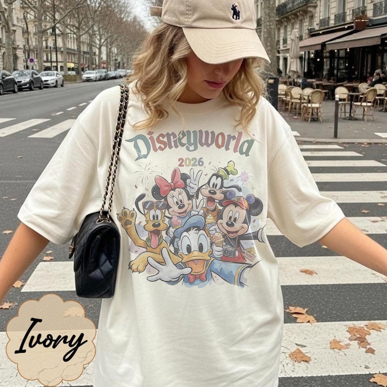 Pu&ograve; includere: T-shirt color avorio con grafica Disneyworld con Topolino, Minnie, Paperino e altri personaggi. Il testo "Disneyworld 2026" &egrave; stampato sopra i personaggi. La maglietta &egrave; abbinata a un berretto da baseball beige e una borsa nera.