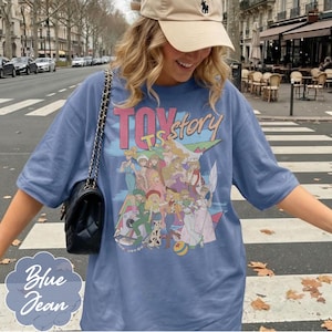 May include: A blue short-sleeved t-shirt featuring a graphic print with the text "TOY story" and an illustration of various female characters in colorful outfits. The shirt is paired with a black quilted shoulder bag with a gold chain strap.