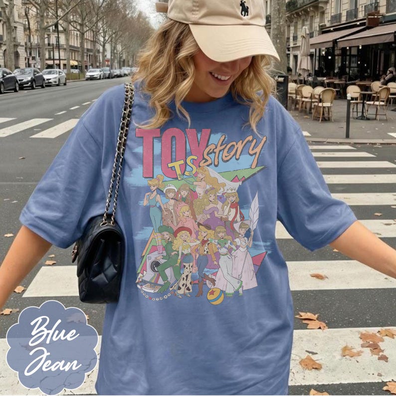 May include: A blue short-sleeved t-shirt featuring a graphic print with the text "TOY story" and an illustration of various female characters in colorful outfits. The shirt is paired with a black quilted shoulder bag with a gold chain strap.
