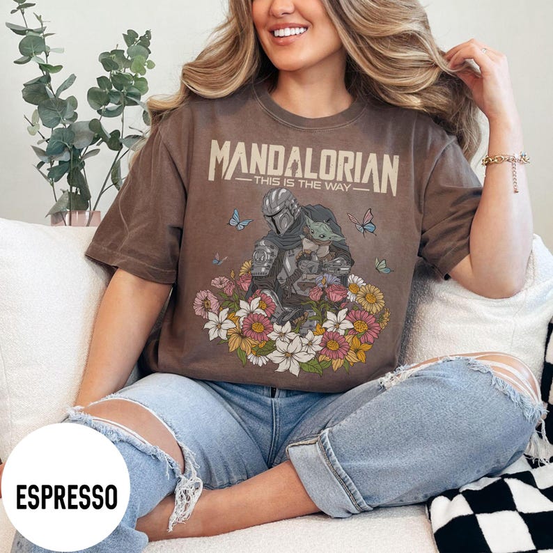 Puede incluir: Camiseta color espresso con un gr&aacute;fico de The Mandalorian y Grogu rodeados de flores y mariposas. El texto "MANDALORIAN - THIS IS THE WAY" est&aacute; impreso en la parte superior. La camiseta es de corte holgado.