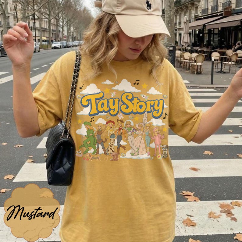 May include: A mustard yellow short-sleeve t-shirt featuring a graphic print with the text Tay Story in blue and yellow letters. The design includes illustrations of various characters, clouds, stars, and musical notes. The shirt has a crew neckline.