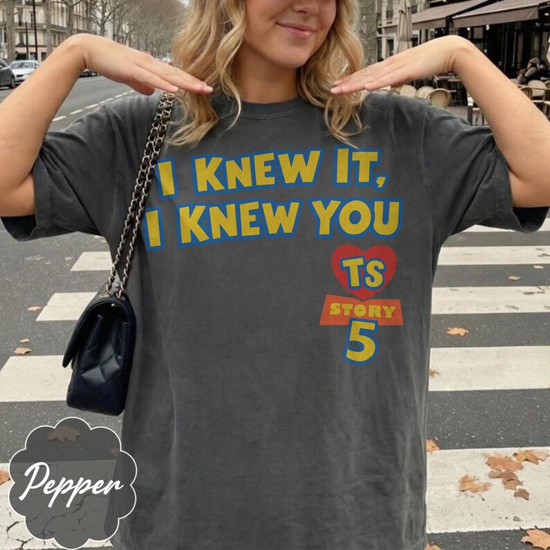 May include: A charcoal gray short-sleeve t-shirt featuring the yellow text I KNEW IT, I KNEW YOU above a red heart with the letters TS, the word STORY, and the number 5 in yellow. A black shoulder bag with a chain strap is worn over the shoulder.