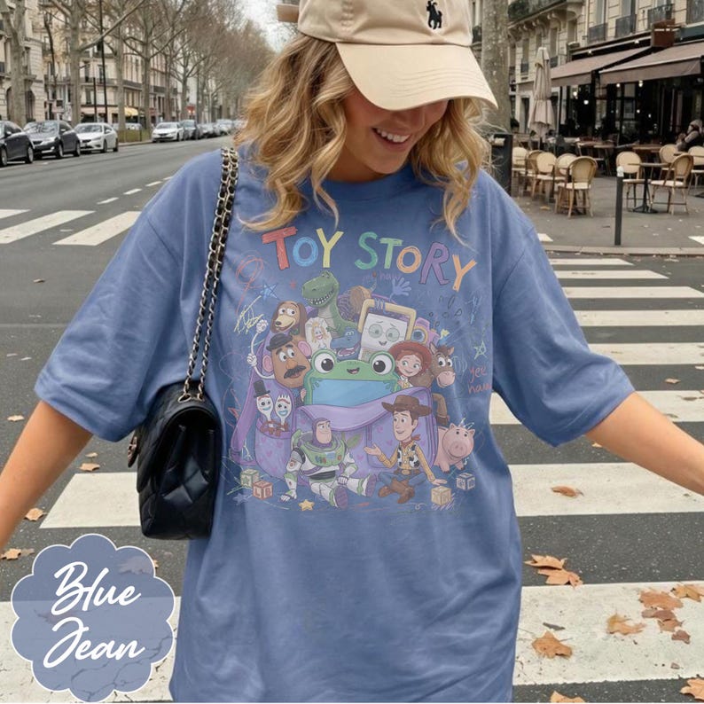 May include: A blue jean coloured t-shirt featuring a graphic print of Toy Story characters including Woody, Buzz Lightyear, and Rex. The text TOY STORY is printed in colourful letters above the characters. A black quilted handbag with a chain strap.