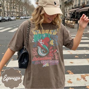 May include: Espresso-colored t-shirt featuring a graphic of Ariel from The Little Mermaid, with the text "Ariel Ocean Tour" and "Sebastian the Crustacean". The shirt has short sleeves and a relaxed fit. A beige cap and black purse are also visible.