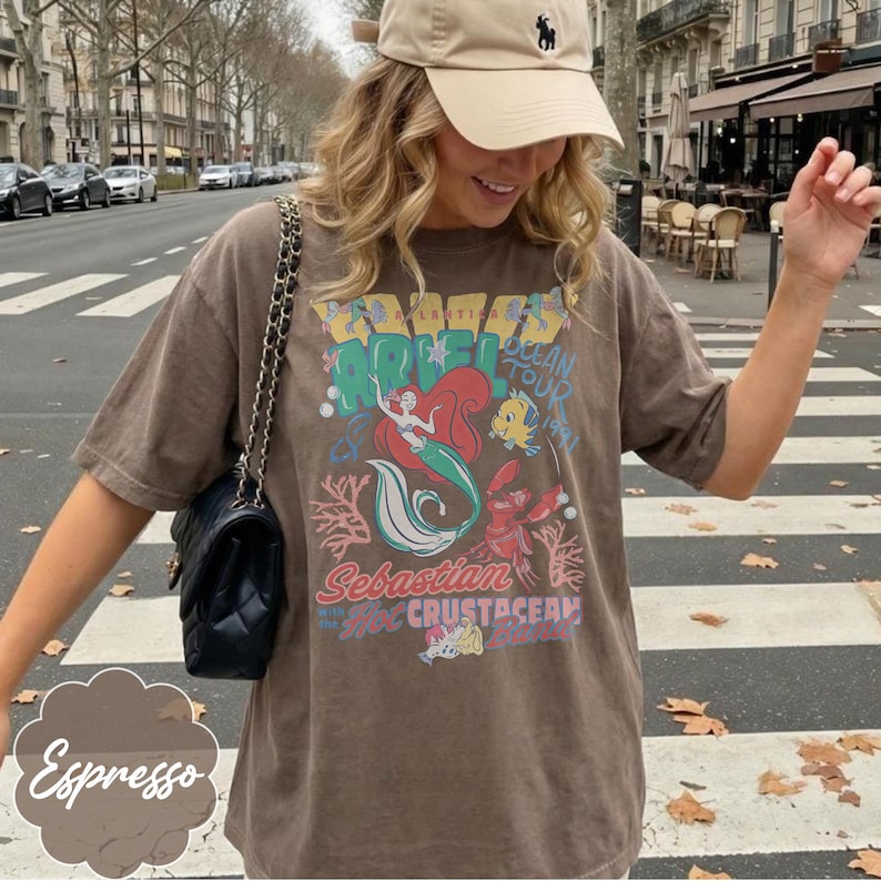 May include: Espresso-colored t-shirt featuring a graphic of Ariel from The Little Mermaid, with the text "Ariel Ocean Tour" and "Sebastian the Crustacean". The shirt has short sleeves and a relaxed fit. A beige cap and black purse are also visible.