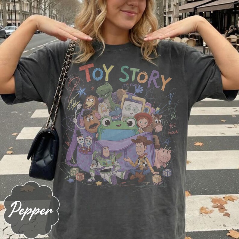 May include: A charcoal grey short-sleeve t-shirt featuring a colourful graphic print of Toy Story characters including Woody, Buzz Lightyear, and Rex. The design includes the text "TOY STORY" in multi-coloured letters and various hand-drawn style doodles.