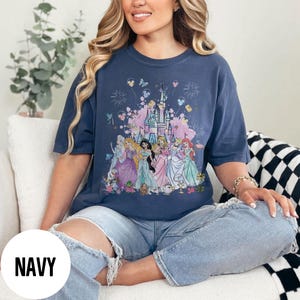May include: Navy blue t-shirt featuring a colorful graphic of Disney princesses in front of a castle. The design includes fireworks, flowers, and balloons. The word "NAVY" is printed on a white circle at the bottom.