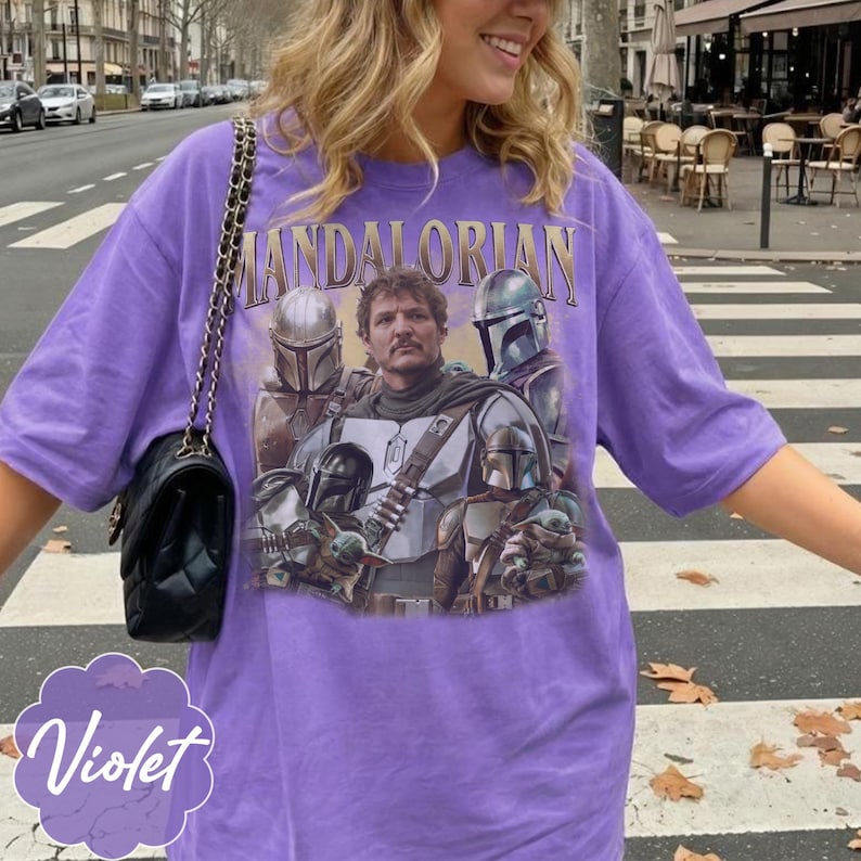 Pu&ograve; includere: Una t-shirt viola a maniche corte con una stampa grafica dei personaggi di The Mandalorian, tra cui una figura centrale, guerrieri in armatura e una piccola creatura. Il testo "MANDALORIAN" &egrave; stampato in un carattere color oro sopra.