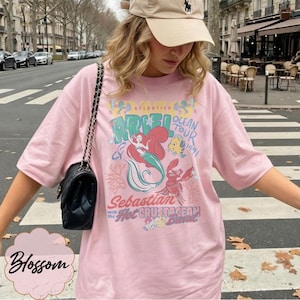 May include: A light pink t-shirt featuring a graphic of Ariel from The Little Mermaid, with the words "Ariel Ocean Tour 1991" and "Sebastian with the Hot Crustacean Band". The shirt has a crew neck and short sleeves.