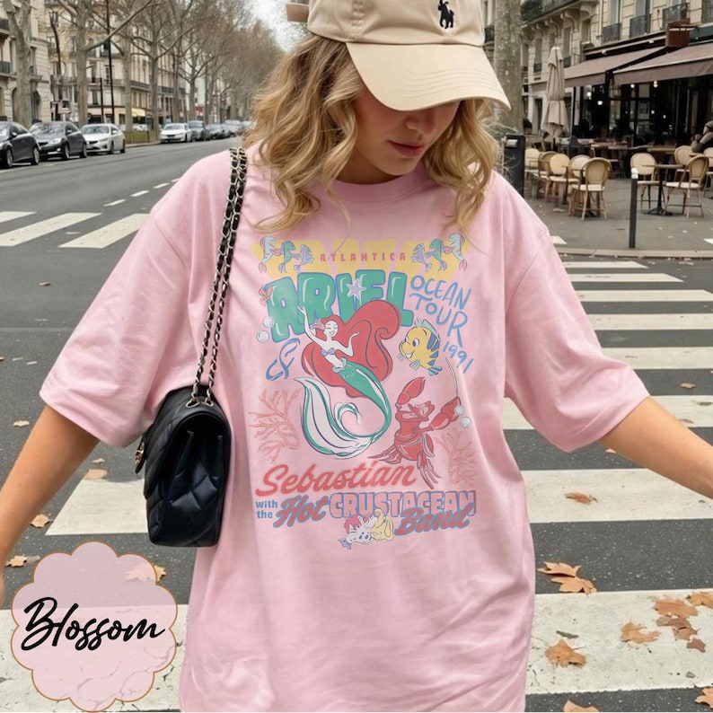 May include: A light pink t-shirt featuring a graphic of Ariel from The Little Mermaid, with the words "Ariel Ocean Tour 1991" and "Sebastian with the Hot Crustacean Band". The shirt has a crew neck and short sleeves.
