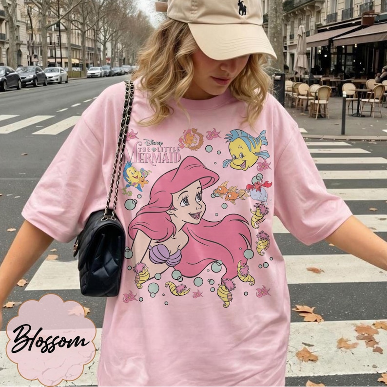 May include: Pale pink t-shirt featuring Ariel from Disney's The Little Mermaid, with Flounder and other characters. The shirt has short sleeves and a crew neck. The text "Disney The Little Mermaid" is printed above Ariel. The word "Blossom" is printed in a cursive font.