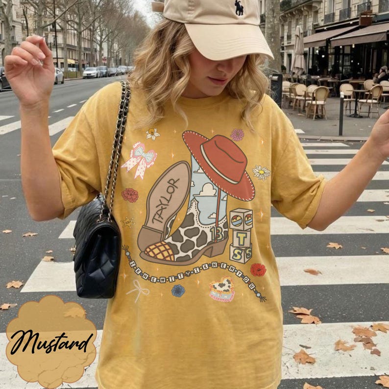 May include: A mustard yellow short-sleeve t-shirt featuring a graphic print of a cowboy boot with a cow print pattern, a red hat, the name Taylor, the number 13, and stacked blocks with the letters T, S, and S. The design includes small floral accents.