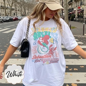 May include: White t-shirt featuring a colorful graphic of Ariel, Sebastian, and Flounder from The Little Mermaid. The design includes the text "Atlantica Ocean Tour 1991" and "Sebastian with the Hot Crustacean Band."