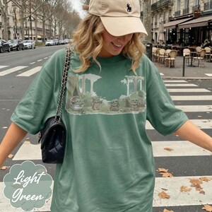 Puede incluir: Camiseta verde claro con un dise&ntilde;o de Grogu de The Mandalorian en una escena de bosque. La camiseta tiene mangas cortas y un corte holgado. Las palabras "Light Green" est&aacute;n impresas en la esquina inferior izquierda.