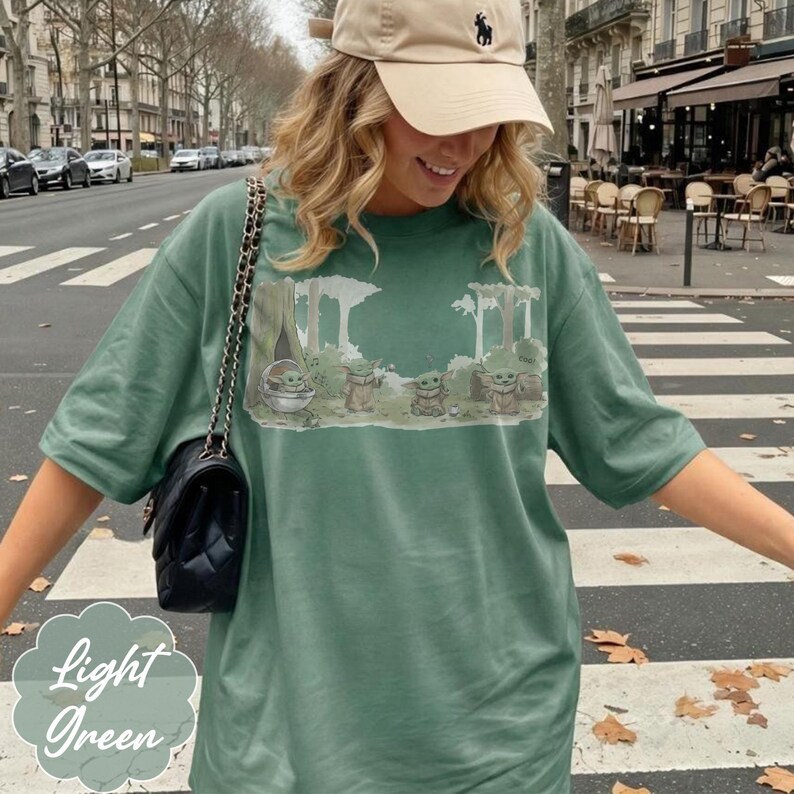 Puede incluir: Camiseta verde claro con un dise&ntilde;o de Grogu de The Mandalorian en una escena de bosque. La camiseta tiene mangas cortas y un corte holgado. Las palabras "Light Green" est&aacute;n impresas en la esquina inferior izquierda.