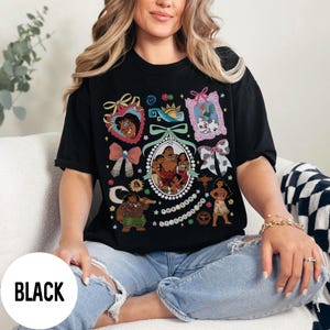 Pu&ograve; includere: T-shirt nera con un design grafico colorato con personaggi ed elementi decorativi. Il design include un ovale centrale con un ritratto di famiglia e varie immagini incorniciate e fiocchi. La parola "BLACK" &egrave; stampata su un cerchio bianco.
