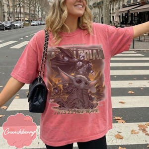 Puede incluir: Una camiseta de manga corta color coral con un estampado gr&aacute;fico de The Mandalorian y Grogu. El texto dice Star Wars The Mandalorian y This Is The Way. La camiseta tiene cuello redondo y un corte holgado, combinada con un bolso de hombro negro acolchado.