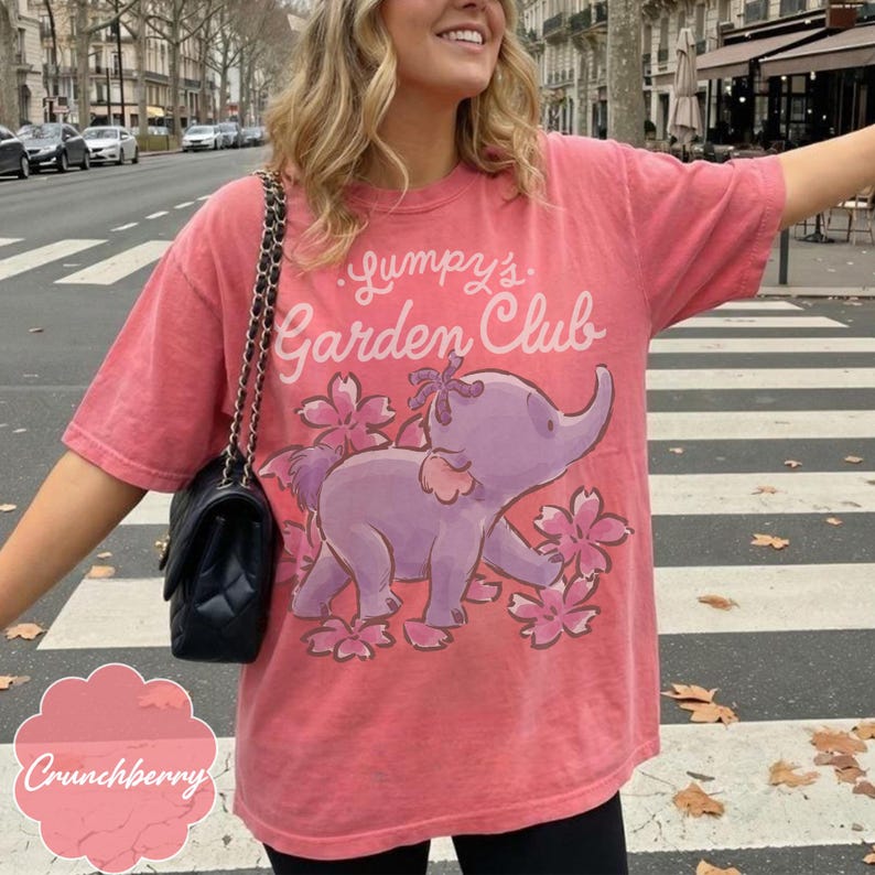 Op de afbeelding: Een koraalkleurig t-shirt met de tekst "Lumpy's Garden Club" boven een paarse olifantafbeelding met roze bloemen. Het shirt heeft korte mouwen en een relaxte pasvorm.
