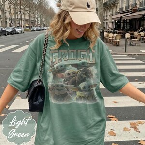 May include: A light green t-shirt featuring a graphic print of multiple Grogu characters and the text GROGU at the top. The shirt has short sleeves and a crew neckline. A black quilted shoulder bag with a gold chain strap is worn over the shoulder.