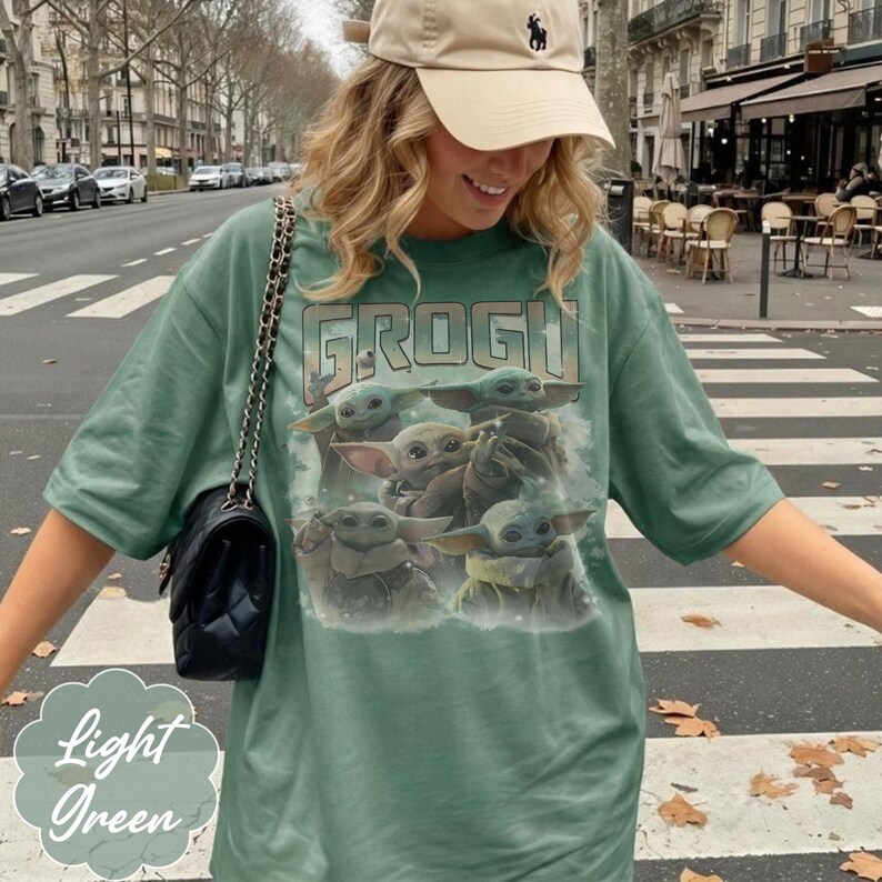 May include: A light green t-shirt featuring a graphic print of multiple Grogu characters and the text GROGU at the top. The shirt has short sleeves and a crew neckline. A black quilted shoulder bag with a gold chain strap is worn over the shoulder.