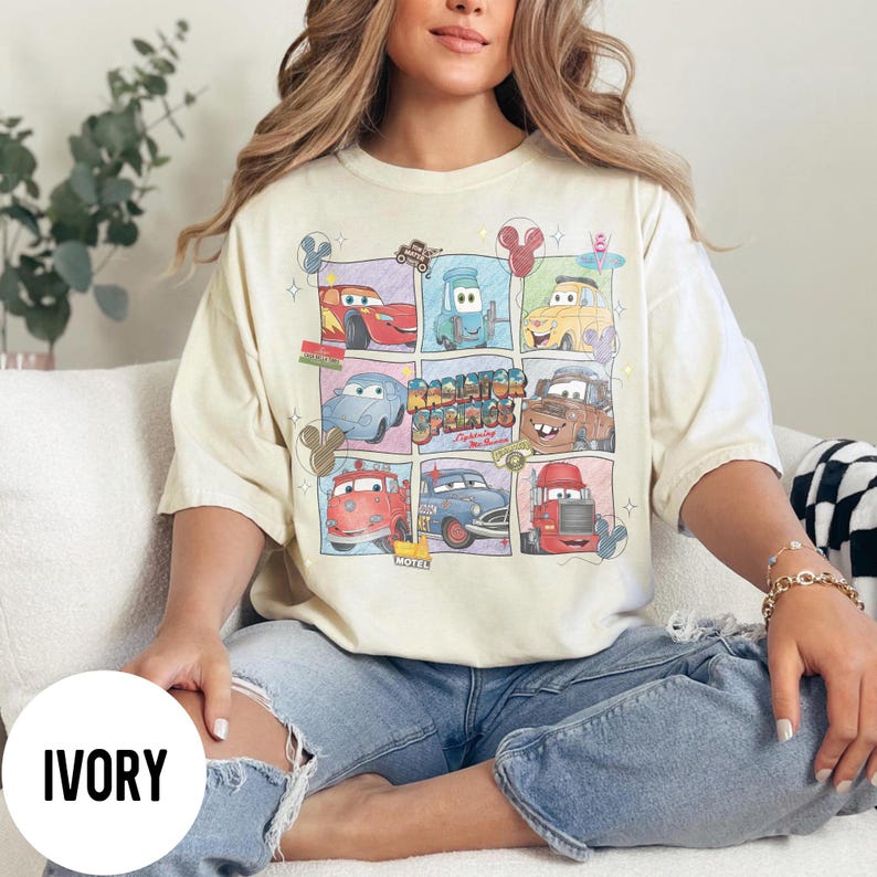 May include: Ivory-colored t-shirt featuring a grid of cartoon cars with the text "Radiator Springs". The design includes various car characters in colorful squares. The shirt is a relaxed fit.