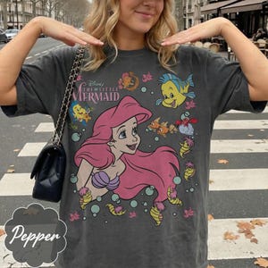 May include: Dark grey t-shirt with a graphic of Ariel from Disney's The Little Mermaid. The design includes Ariel with pink hair, Flounder, and other sea creatures. The shirt has the text "Disney The Little Mermaid" across the top.