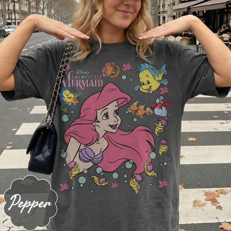May include: Dark grey t-shirt with a graphic of Ariel from Disney's The Little Mermaid. The design includes Ariel with pink hair, Flounder, and other sea creatures. The shirt has the text "Disney The Little Mermaid" across the top.