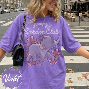 Op de afbeelding: Violetkleurig T-shirt met de tekst "Gumpy's Garden Club" en een cartoon olifantenontwerp. De olifant is paars met roze bloemaccenten. Het woord "Violet" is in een gestileerd lettertype linksonder geschreven.