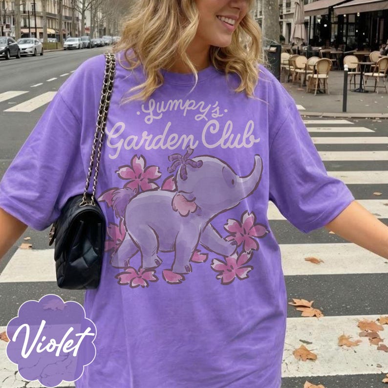 Op de afbeelding: Violetkleurig T-shirt met de tekst "Gumpy's Garden Club" en een cartoon olifantenontwerp. De olifant is paars met roze bloemaccenten. Het woord "Violet" is in een gestileerd lettertype linksonder geschreven.