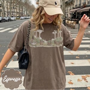 Puede incluir: Camiseta color espresso con un gr&aacute;fico de Grogu de The Mandalorian. El dise&ntilde;o muestra a Grogu en un bosque con otros personajes. La camiseta se combina con una gorra beige y un bolso negro con correa de cadena.