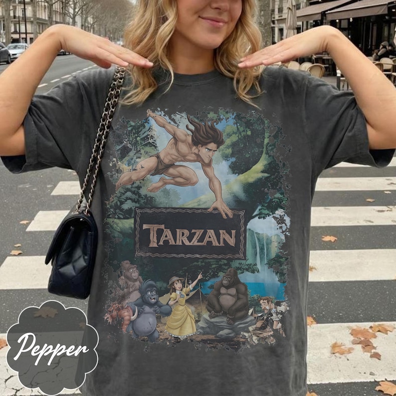 Op de afbeelding: Donkergrijs Tarzan grafisch t-shirt met Tarzan in een junglesc&egrave;ne met het woord "Tarzan" in een rechthoekig kader. Het shirt heeft ook stripfiguren. Het shirt is een vintage stijl.