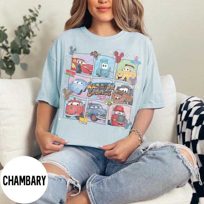 May include: Light blue t-shirt featuring a colorful graphic of cartoon cars in a grid pattern. The design includes the text "Radiator Springs" and various car characters. The shirt is a relaxed fit.