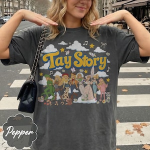 May include: A charcoal gray t-shirt featuring a graphic print with the text "Tay Story" in yellow letters. The design includes illustrations of various characters in different outfits, surrounded by white clouds, yellow stars, and musical notes.