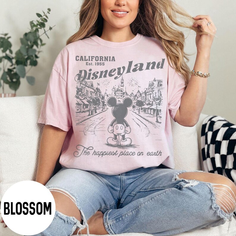 Może przedstawiać: Jasnor&oacute;żowa koszulka z grafiką Disneyland z Myszka Miki. Grafika zawiera tekst "CALIFORNIA Est. 1955 Disneyland" i "The happiest place on earth". Koszulka jest noszona przez osobę.