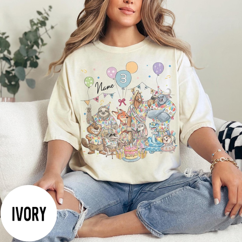 以下が含まれることがあります： アイボリー色のTシャツ。漫画の動物、風船、ケーキが描かれた、カラフルな誕生日のパーティーシーンが特徴です。「Name」と数字の「3」の文字も表示されています。このシャツは快適でカジュアルなスタイルです。