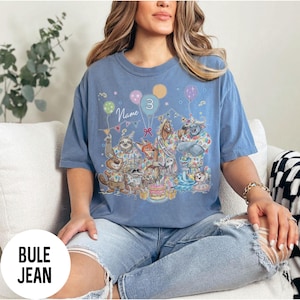 以下が含まれることがあります： 誕生日を祝う擬人化された動物たちのカラフルなイラストが描かれた青いTシャツ。グラフィックには風船、ケーキ、数字の「3」が含まれています。「Name」という言葉も表示されています。