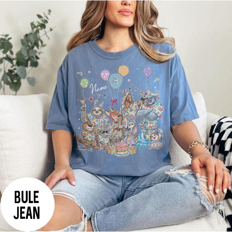 以下が含まれることがあります： 誕生日を祝う擬人化された動物たちのカラフルなイラストが描かれた青いTシャツ。グラフィックには風船、ケーキ、数字の「3」が含まれています。「Name」という言葉も表示されています。