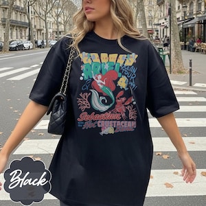May include: Black t-shirt featuring a colorful graphic of Ariel, Sebastian, and Flounder from The Little Mermaid. The design includes the text "Ariel Ocean Tour 1991" and "Sebastian with the Hot Crustacean Band."