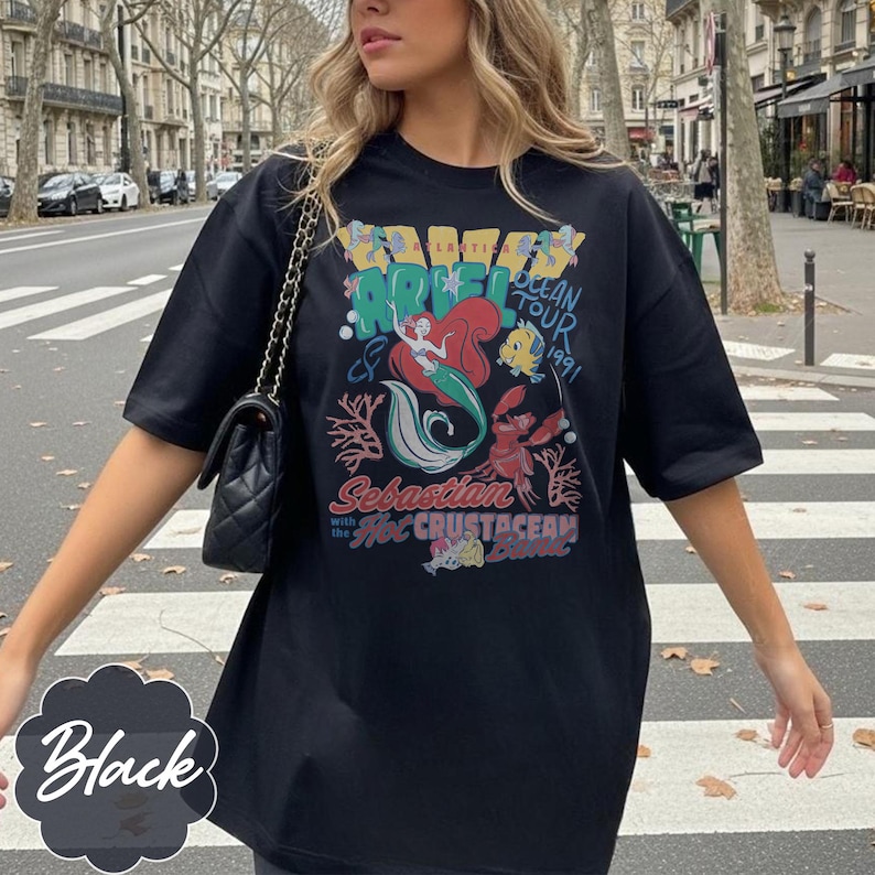 May include: Black t-shirt featuring a colorful graphic of Ariel, Sebastian, and Flounder from The Little Mermaid. The design includes the text "Ariel Ocean Tour 1991" and "Sebastian with the Hot Crustacean Band."