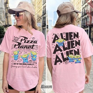 K&ouml;nnte beinhalten: Hellrosa T-Shirt mit dem Pizza Planet Logo und Alien-Charakteren. Auf der Vorderseite des Shirts steht "Pizza Planet". Auf der R&uuml;ckseite des Shirts steht das Wort "Alien" wiederholt mit Alien-Grafiken.