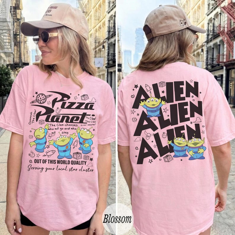 K&ouml;nnte beinhalten: Hellrosa T-Shirt mit dem Pizza Planet Logo und Alien-Charakteren. Auf der Vorderseite des Shirts steht "Pizza Planet". Auf der R&uuml;ckseite des Shirts steht das Wort "Alien" wiederholt mit Alien-Grafiken.