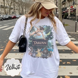 Op de afbeelding: Wit T-shirt met een Tarzan-afbeelding. Het ontwerp toont Tarzan in een junglesc&egrave;ne, met het woord "Tarzan" in bruin. Het shirt heeft korte mouwen en een ronde hals.