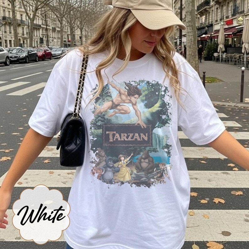Op de afbeelding: Wit T-shirt met een Tarzan-afbeelding. Het ontwerp toont Tarzan in een junglesc&egrave;ne, met het woord "Tarzan" in bruin. Het shirt heeft korte mouwen en een ronde hals.